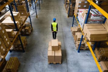 Warehouse worker in warehouse with yellow vest and blue hardhat pulling a cart with boxes
